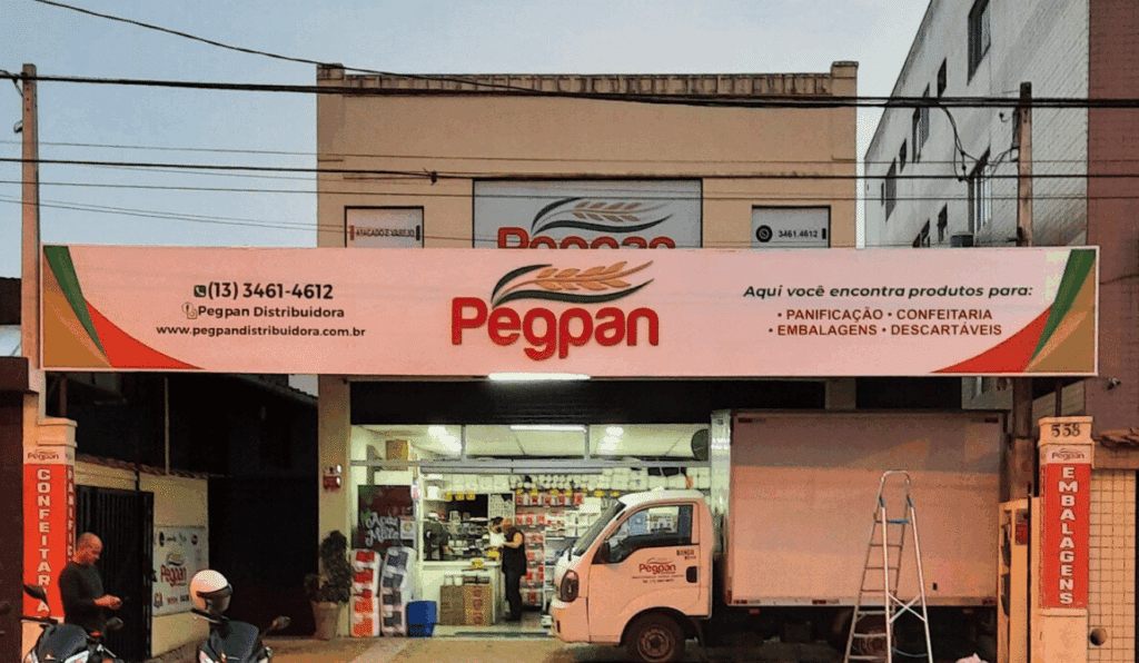 Fachada em ACM na cor branca com logotipo em PVC Expandido colorido de acordo com as cores da marca, iluminada por refletores. A empresa fica localizada na cidade de São Vicente-SP.